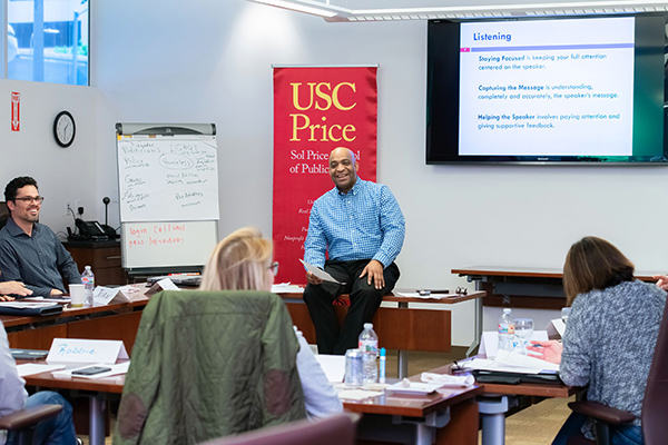 Darly Dixon in a blue shirt sitting in front of the classroom smiling in front of USC Price banner