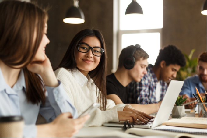 Girl with glasses smiling and a fellow student in a study hall full of young students