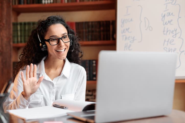 A lady wearing glasses and wireless headest at virtual meeting, sitting at desk, having video call on laptop, waving to webcam. Woman studying or teaching online at home.
