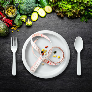 Measuring tape and pills are on a white plate with vegetables on the table above the plate