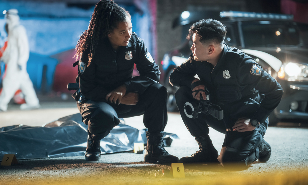 Two police officers crouch and talk to each other at a crime scene