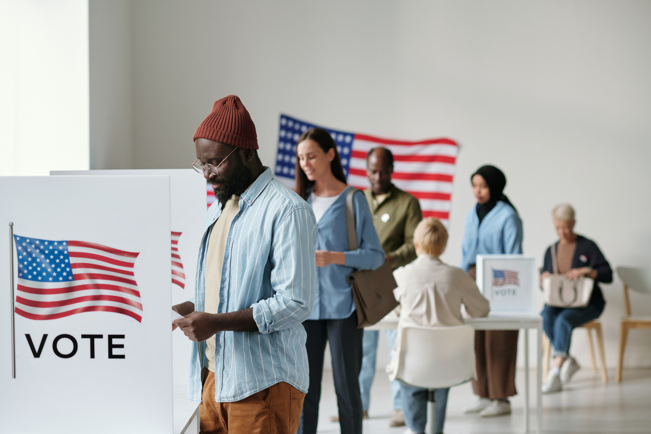 Group of young voters in casualwear standing in queue along vote booths in polling place and putting their ballots into boxes