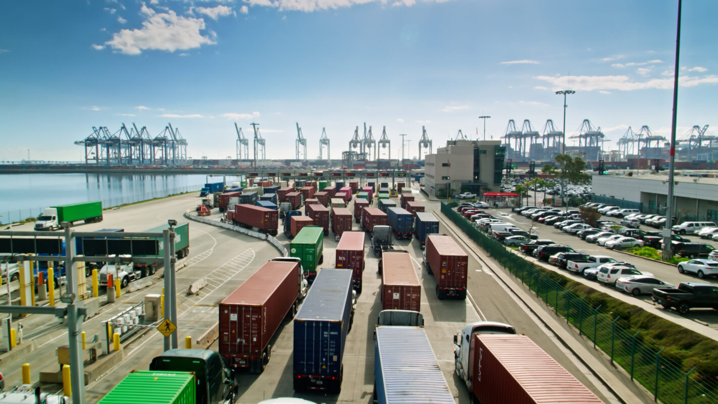 Drone shot of the front gate of a container terminal in the Port of Los Angeles on a sunny day. Trucks are lined up to leave and enter, and in the background massive container ships are sitting in their berths being loaded and unloaded by cranes. Authorization was obtained from the Los Angeles Harbor Department and Los Angeles Port Police for this operation in restricted airspace.