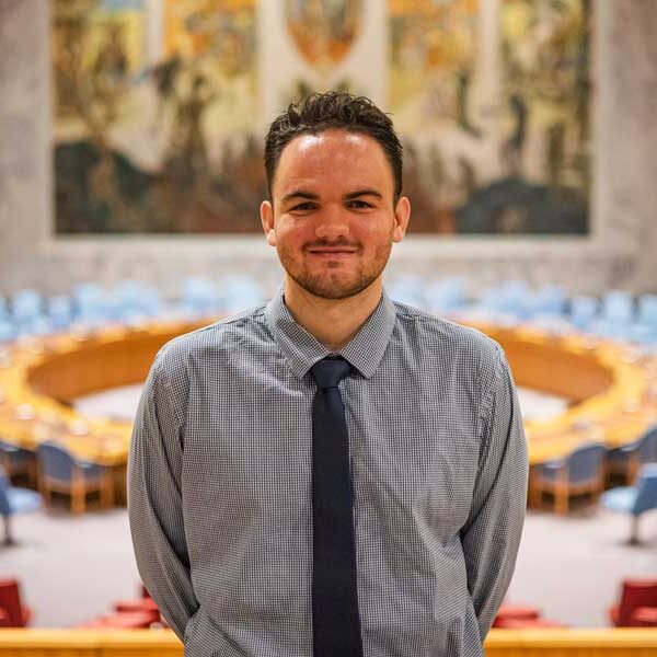 Elijah Dow poses for a photo at the United Nations Security Council Chamber wearing a grey shirt and dark blue tie