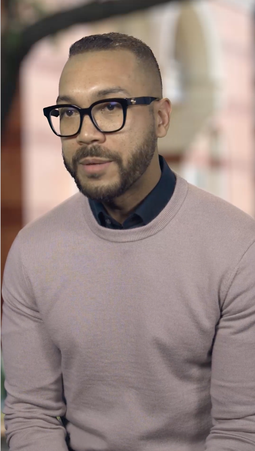 Man wearing black-rimmed glasses in a mauve sweater vest.