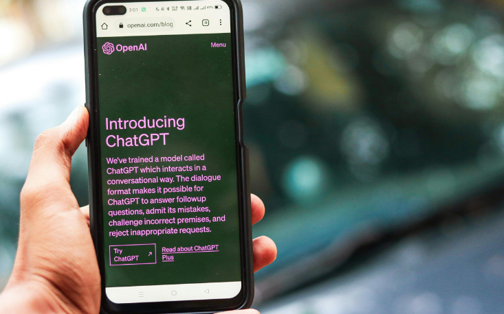A hand holding a smartphone displaying the OpenAI blog page introducing ChatGPT, with text describing its conversational abilities illustrating the integration of AI in Higher Education through accessible, interactive technology.
