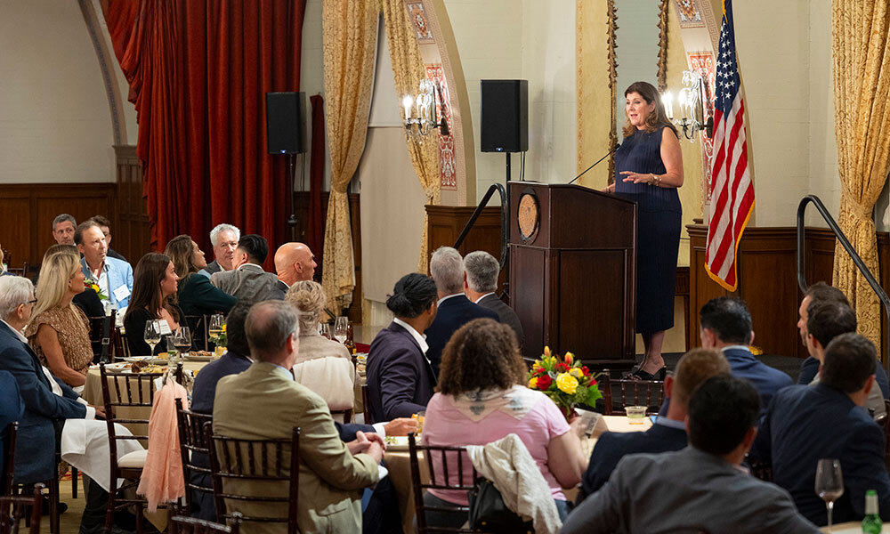 Attendees at the Chandler Dinner sit at tables while Clare De Briere speaks on stage behind a podium.