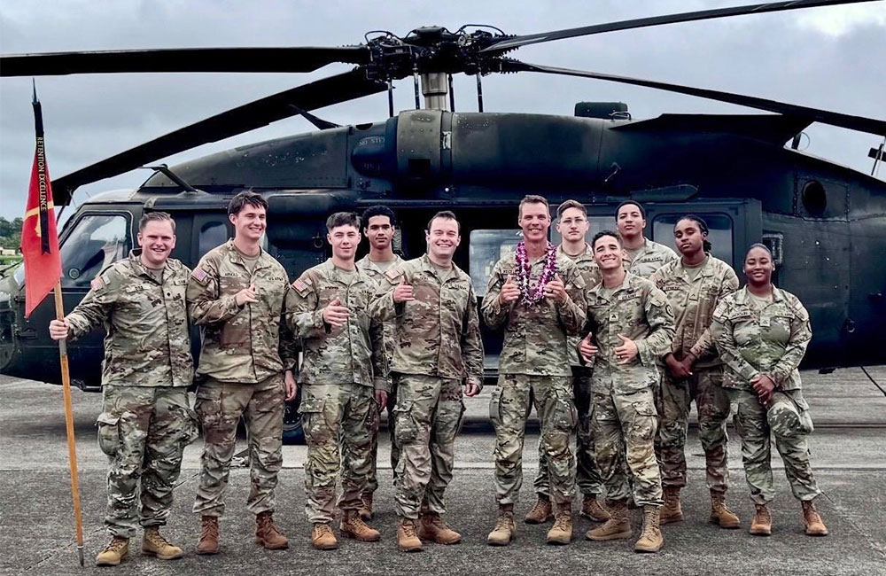 Eleven U.S. Army soldiers in camouflage uniforms pose together in front of a Black Hawk helicopter on a tarmac, with one soldier holding a guidon and another wearing a flower lei.